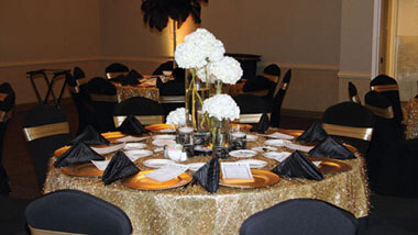 Blue chairs surround a circular table covered with a gold table cloth, gold plates and black napkins. A centerpiece contains white flowers.
